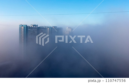 A white-and-blue multi-storey residential building is shrouded in fog in the early spring morning. 110747556