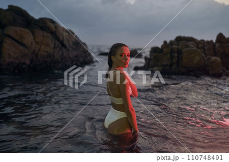 Attractive woman enjoys nighttime ocean swim illuminated by crimson light, elegance in aquatic setting. Female basks in sea glow, fashion swimwear visible, night beach leisure scene captures allure. 110748491