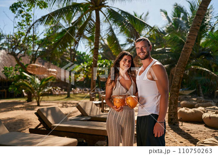 Smiling couple enjoys honeymoon in tropical paradise, sharing fresh coconut water. Fit man and woman in summer attire stand amidst palm trees, celebrating romantic getaway with exotic drinks. 110750263