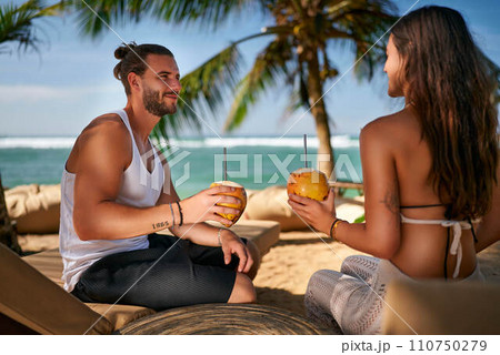 Couple enjoys fresh coconut water on tropical beach. Man, woman sip cocktails, relax under palm trees by sea. Romantic getaway, leisure on sandy shore. Vacationers drink, bask in summer sun, luxuriate 110750279