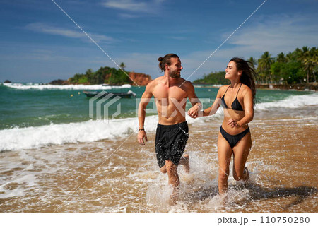 Joyful couple runs on tropical beach. Fit man and slender woman enjoy active vacation, sprinting by sea. Athletic duo practices beach running, promotes wellness. Excited pair in swimwear share fun. 110750280