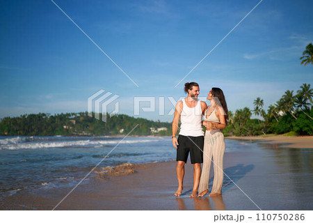 Romantic couple strolls on beach, holding hands by the sea. Man, woman enjoy honeymoon at tropical coast. Casual beachwear, sunset walk, love, vacation. Smiling duo in relaxed seaside promenade. 110750286