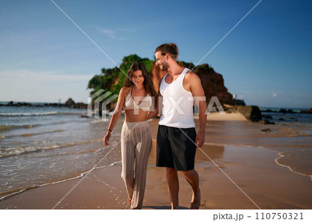 Romantic couple strolls on beach at sunset, holding hands. Woman, man walk barefoot on sand by sea, enjoy vacation. Trendy, young lovers on seaside date. Relaxed, carefree tourists on coastal escape. Romantic couple strolls on beach at sunset, holding hands. Woman, man walk barefoot on sand by sea, enjoy vacation. Trendy, young lovers on seaside date. Relaxed, carefree tourists on coastal escape. 110750321