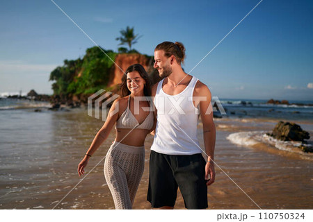 Romantic couple strolls on tropical beach at sunset. Newlyweds on honeymoon enjoy seaside walk. Man, woman in casual beachwear, bonding, smiling, love. Ocean, vacation, travel, summer, love concept. Romantic couple strolls on tropical beach at sunset. Newlyweds on honeymoon enjoy seaside walk. Man, woman in casual beachwear, bonding, smiling, love. Ocean, vacation, travel, summer, love concept. 110750324