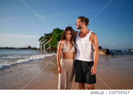 Couple walks on beach, adult duo sans kids enjoys seaside stroll. Childless pair on vacation smile, unwind by ocean, travel trends. Man, woman in summer attire embrace leisure, tropical getaway. 110750327