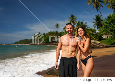Fit couple stands on tropical beach, athletic man and slender woman smile, enjoying eco travel. Abs, attractive duo on vacation, seaside workout, sunny coastal lifestyle, healthy living concept. Fit couple stands on tropical beach, athletic man and slender woman smile, enjoying eco travel. Abs, attractive duo on vacation, seaside workout, sunny coastal lifestyle, healthy living concept. 110750369
