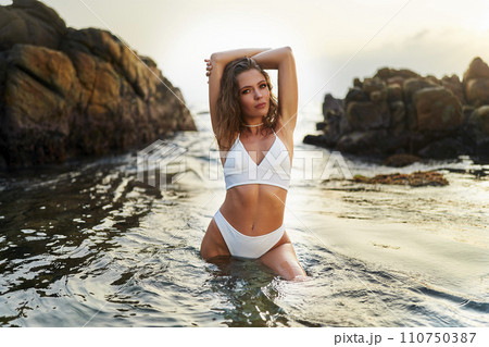 Attractive woman in stylish white two-piece posing in ocean water near rocks during sunset. Fashionable female in chic white bikini stands in sea, sunset light glistens on water around her. 110750387
