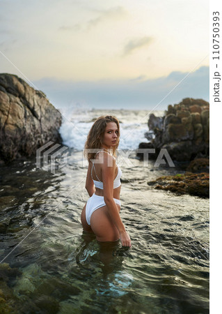 Woman in white swimsuit poses in sea water, fashion model explores tropical coastline, travel destination beauty shot. 110750393