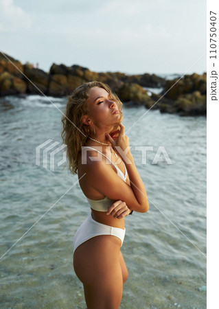 Young woman enjoying sun at seaside, wearing swimsuit, coastal rocks backdrop, serenity concept 110750407
