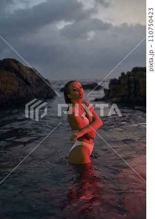 Attractive woman enjoys a serene night swim in the ocean, bathed in a red glow, rocks nearby, travel and leisure concept, nocturnal beach activity, beauty in nature, peaceful getaway. 110750423