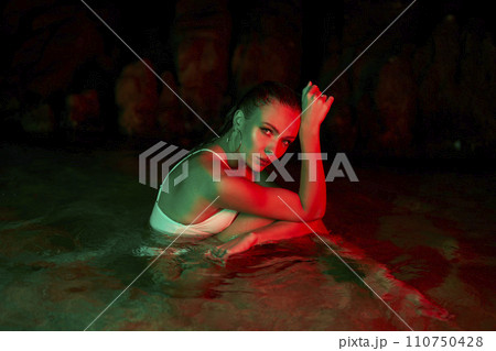 Woman sits in water at night, illuminated by red and green lights, expressing contemplation and moodiness, wet hair, focused gaze, feeling of solitude. Woman sits in water at night, illuminated by red and green lights, expressing contemplation and moodiness, wet hair, focused gaze, feeling of solitude. 110750428