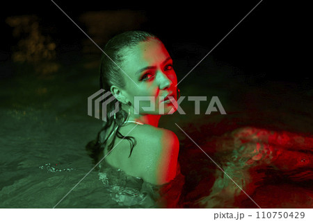 Woman immersed in lagoon at night, lit by green and red hues, reflects beauty, trend, and elegance, during a nocturnal water photo shoot, showcasing fashion amidst natural elements. 110750429
