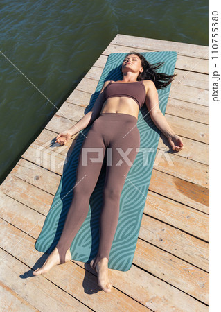 A slender woman practicing yoga, doing a relaxing shavasana exercise after the main workout, lies in sportswear on a mat near the lake 110755380