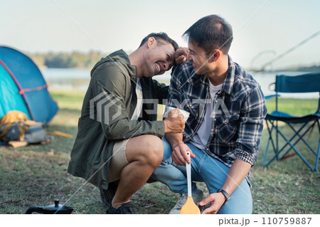 Gay LGBTQIA couple sitting on picnic chair drinking tea and making food while camping on vacation holiday Gay LGBTQIA couple sitting on picnic chair drinking tea and making food while camping on vacation holiday 110759887