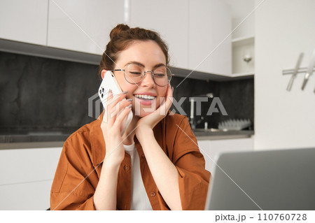 Portrait of beautiful young woman managing her own business from home, freelancer making phone call, sitting in kitchen with laptop and smiling Portrait of beautiful young woman managing her own business from home, freelancer making phone call, sitting in kitchen with laptop and smiling 110760728