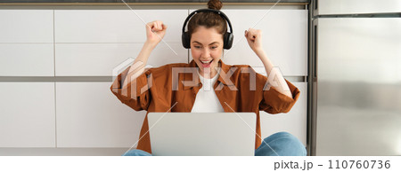 Excited young woman, programmer achieves goal, triumphing, winning on laptop, sitting in headphones, dancing with hands, celebrating victory 110760736