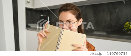 Close up portrait of smiling beautiful woman with notebook, making notes, writing in diary, studying at home, preparing homework at in kitchen 110760755