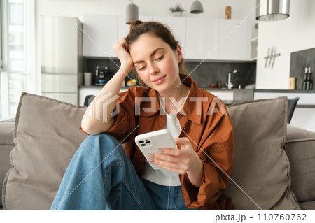 Portrait of young modern woman reading on mobile phone, scrolling social media app on smartphone, sitting on couch in living room in casual clothes 110760762