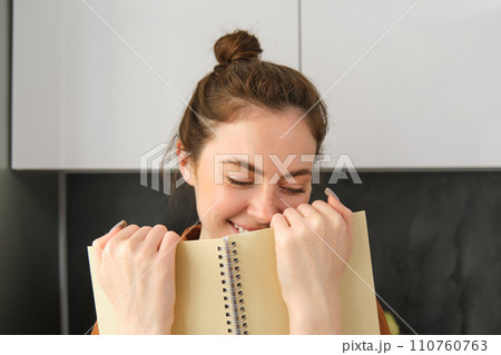 Coquettish young woman smiling, hiding face behind notebook, laughing and smiling, standing in the kitchen Coquettish young woman smiling, hiding face behind notebook, laughing and smiling, standing in the kitchen 110760763