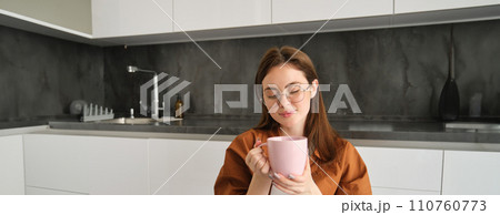 Relaxing day at home. Smiling young woman in glasses, enjoying fresh brewed cup of coffee, holding mug and sitting in kitchen, resting on peaceful autumn day at home Relaxing day at home. Smiling young woman in glasses, enjoying fresh brewed cup of coffee, holding mug and sitting in kitchen, resting on peaceful autumn day at home 110760773