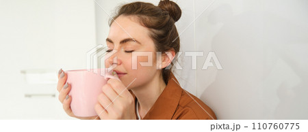 Close up portrait of beautiful brunette woman with cup of coffee, drinking aromatic beverage from mug, smiling from pleasure, sitting in kitchen at home Close up portrait of beautiful brunette woman with cup of coffee, drinking aromatic beverage from mug, smiling from pleasure, sitting in kitchen at home 110760775