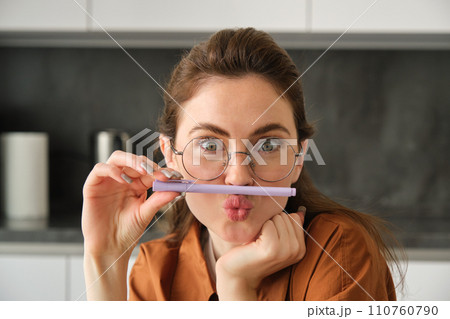 Close up portrait of funny, silly young woman, playing with pen, holding pencil on top of her lip and grimacing, sitting in kitchen 110760790