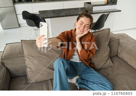Portrait of beautiful smiling woman, takes selfie at home, posing on sofa, holds smartphone with extended hand 110760805