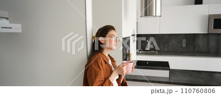 Portrait of happy young woman at home, holding cup of tea, leaning on wall, smiling, drinking coffee from pink mug 110760806