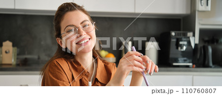 Portrait of beautiful female student in glasses, has online lesson, sits at home, holds pen, doing homework, smiling and looking at camera 110760807