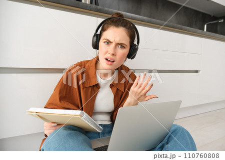 Portrait of annoyed and confused woman in headphones, sits on floor with laptop and notebook, looks frustrated, has issues in doing homework or studying 110760808