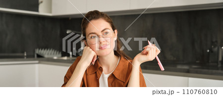 Close up of dreamy smiling woman, sitting in kitchen with pen, looking outside window, writing, making notes Close up of dreamy smiling woman, sitting in kitchen with pen, looking outside window, writing, making notes 110760810