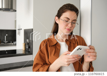 Portrait of young woman at home, checking news or messages on smartphone, online shopping, confirm delivery on mobile phone app, leaning on wall, standing in living room 110760815