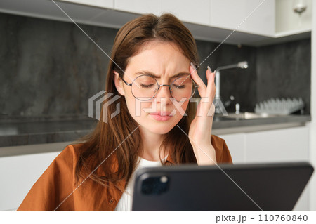 Close up portrait of brunette woman in glasses, grimacing from painful migraine or headache, feels unwell while reads digital tablet, works from home in kitchen Close up portrait of brunette woman in glasses, grimacing from painful migraine or headache, feels unwell while reads digital tablet, works from home in kitchen 110760840
