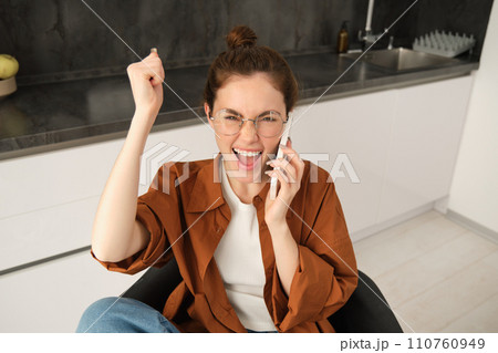 Portrait of excited woman in kitchen, sitting with mobile phone, celebrating, receives great news over the telephone, triumphing 110760949