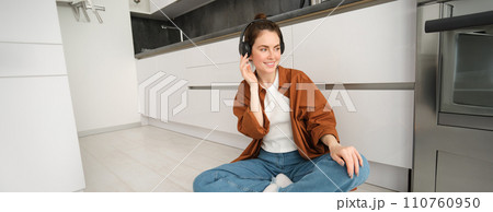 Stylish modern woman in wireless headphones, sits on floor at home, listens to music or podcast, enjoys sound quality in her brand new earphones 110760950