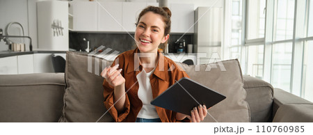 Portrait of smiling, carefree brunette woman, sitting on couch in living room, online shopping or reading on digital tablet, looking happy at camera 110760985