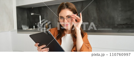 Portrait of beautiful young female model in glasses, holding digital tablet, wearing glasses, reading e-book, posing in kitchen at home 110760997