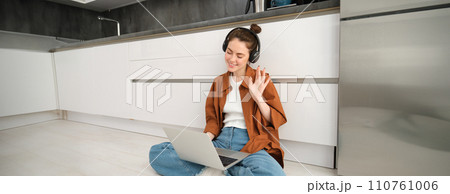 Portrait of happy woman in headphones, looking at laptop, waving hand at computer, chatting on video online, joins team meeting, calling friend on computer, sitting on floor 110761006