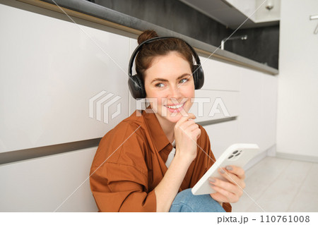 Positive young woman, sits at home with wireless headphones and smartphone, smiles and looks happy Positive young woman, sits at home with wireless headphones and smartphone, smiles and looks happy 110761008