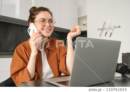 Portrait of young self-employed woman, entrepreneur working from home, freelancer calling client. Girl making an order, talking to someone on phone, sitting in kitchen with laptop Portrait of young self-employed woman, entrepreneur working from home, freelancer calling client. Girl making an order, talking to someone on phone, sitting in kitchen with laptop 110761033