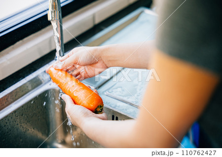 A young woman meticulously washes carrots in the kitchen sink emphasizing hygiene and organic food preparation. Illustrating the concept of washing vegetables before cooking. A young woman meticulously washes carrots in the kitchen sink emphasizing hygiene and organic food preparation. Illustrating the concept of washing vegetables before cooking. 110762427