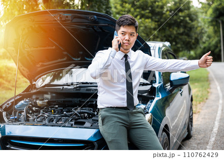 Asian businessman stranded on the road after car breakdown, using phone to call for help. Candid shot of man waiting for assistance and giving thumbs up. Transportation and insurance concept. 110762429