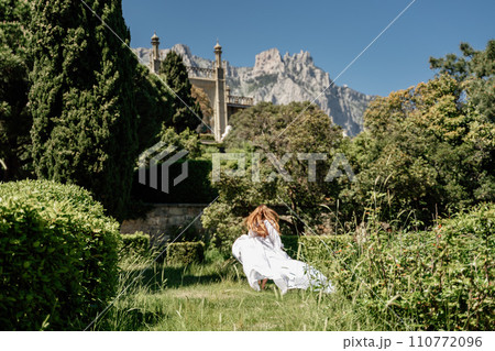 a beautiful woman with long brown hair and long white dress stay along a path along beautiful bushes in the park rear view 110772096