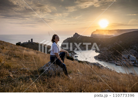 Happy woman on sunset in mountains. Woman siting with her back on the sunset in nature in summer. Silhouette. 110773396