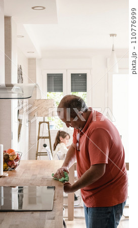 Man cleaning the surface of a kitchen while his granddaughter playing Man cleaning the surface of a kitchen while his granddaughter playing 110776999