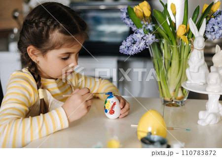 Cute four year girl in beige apron sitting on kitchen table and painting easter eggs Cute four year girl in beige apron sitting on kitchen table and painting easter eggs 110783872