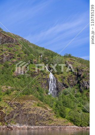 Vertical view of a waterfall through greenery against a blue sky in Trollfjorden 110785299