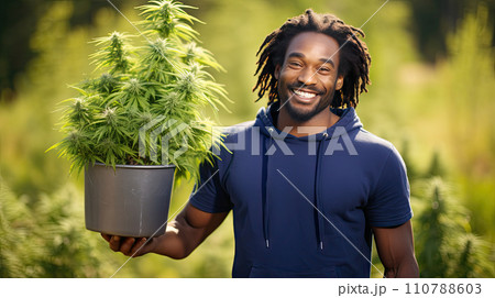 A man holds a pot of flowering cannabis against a green field. 110788603