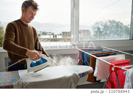 Mature man ironing clothes at home. The concept of maintaining cleanliness and taking care of health. Mature man ironing clothes at home. The concept of maintaining cleanliness and taking care of health. 110788943