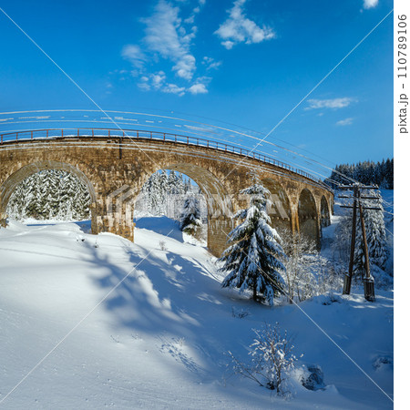 Stone viaduct (arch bridge) on railway through mountain snowy fir forest. Snow drifts  on wayside and hoarfrost on trees and electric line wires. 110789106